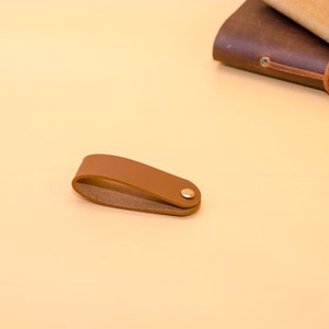 May include: A brown leather bookmark with a gold metal button. The bookmark is on a light brown surface.