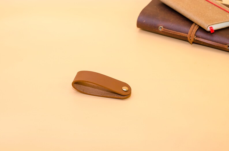May include: A brown leather bookmark with a gold metal button. The bookmark is on a light brown surface.