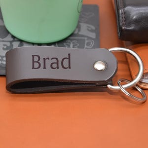 Könnte beinhalten: Ein brauner Leder-Schlüsselanhänger mit einem silbernen Schlüsselring. Der Schlüsselanhänger trägt den Namen "Brad" eingraviert.