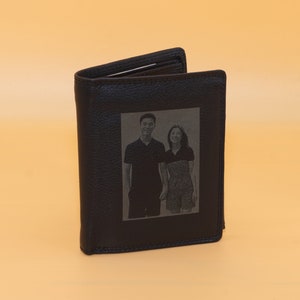 May include: Black leather wallet with a rectangular photo of a couple engraved on the front. The wallet is open, revealing the interior. The background is a solid, light orange color.