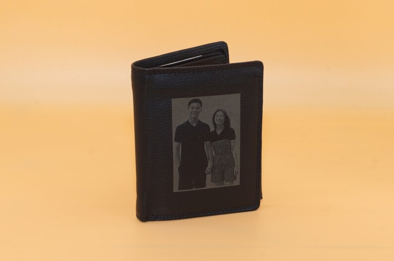 May include: Black leather wallet with a rectangular photo of a couple engraved on the front. The wallet is open, revealing the interior. The background is a solid, light orange color.