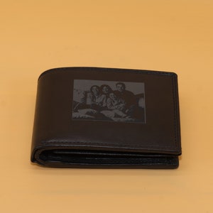May include: A black leather wallet with a rectangular image of a family etched on the front. The wallet is folded and has a classic bi-fold design. The background is a solid light orange.