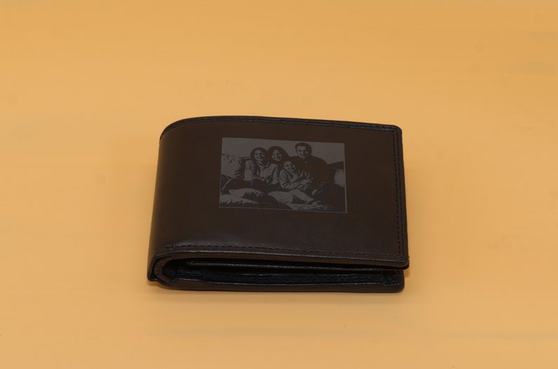 May include: A black leather wallet with a rectangular image of a family etched on the front. The wallet is folded and has a classic bi-fold design. The background is a solid light orange.