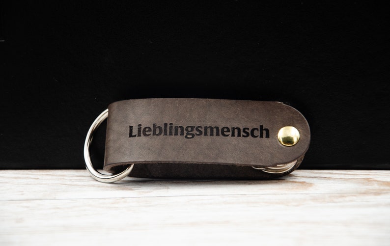May include: A brown leather keychain with a silver ring and a gold button. The keychain is engraved with the text "Lieblingsmensch".