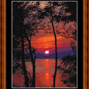 Puede incluir: Obra de arte en punto de cruz enmarcada que representa una puesta de sol sobre el agua. La escena presenta un cielo naranja y rojo vibrante, un sol brillante y la silueta de árboles. La obra está enmarcada en un marco de madera marrón.