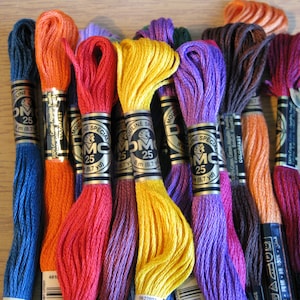 May include: A collection of 10 spools of DMC embroidery floss in various colors, including blue, orange, red, yellow, purple, brown, and pink. Each spool is labeled with the DMC brand name and the number 25, indicating the weight of the floss. The spools are arranged in a row on a wooden surface.