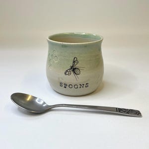 May include: A ceramic honey pot with a green and white glaze. The pot has a bee design and the word "SPOONS" written on the front. A silver spoon is next to the pot.