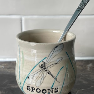 May include: A beige ceramic spoon holder featuring a dragonfly illustration and the word "SPOONS". The holder has blue and green accents, and a silver spoon is placed inside.