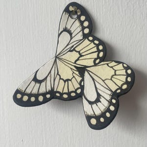 May include: A wall-mounted butterfly ornament crafted from a flat material, featuring a cream-coloured surface with bold black outlines and intricate wing patterns. The butterfly has a small circular hole at the top for hanging and a matte finish.