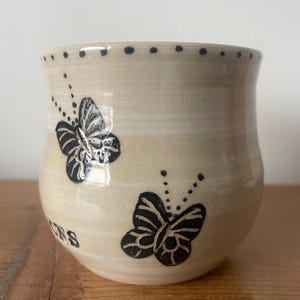 May include: A beige ceramic mug featuring two black butterfly designs and the word "ONS". The mug has a dotted pattern around the rim. The butterflies are detailed with black lines and dots.