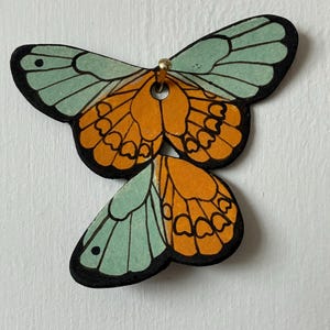 May include: A flat, butterfly-shaped decorative object featuring a colour palette of mint green and vibrant orange. The wings are outlined in black with detailed vein patterns and a small circular hole at the top centre secured by a gold-toned pin.