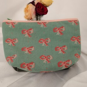 May include: A green fabric pouch with a pink zipper and a pattern of pink and white bows.
