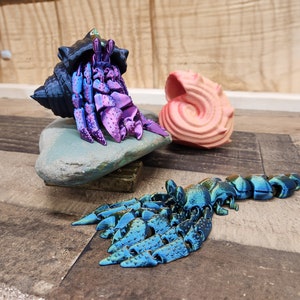 May include: A 3D-printed hermit crab with a blue and purple body, emerging from a dark blue shell. A pink seashell sits nearby. The crab rests on a stone, with a blue and green articulated tail extending outward.