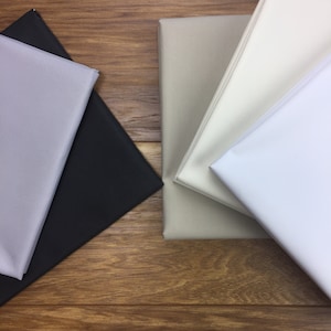 Puede incluir: Cuatro muestras de tela en diferentes colores: gris claro, negro, beige y blanco. Las muestras están dispuestas en forma de cuadrado sobre una superficie de madera.