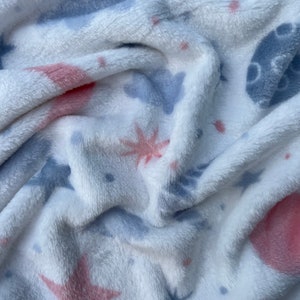 May include: White plush blanket featuring a blue and pink star design. The blanket is soft and fluffy, ideal for snuggling up on the sofa.