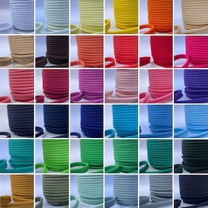 Op de afbeelding: Een verzameling spoelen met kleurrijke biesband in diverse tinten, waaronder wit, geel, oranje, rood, roze, paars, blauw, groen en bruin. Elke spoel is netjes opgerold, met een voorbeeld van de bies.