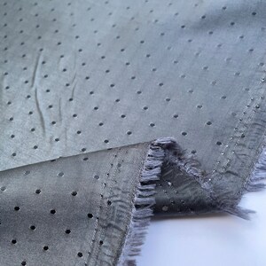 May include: A close-up shot of grey fabric featuring small, evenly spaced holes. The fabric is slightly creased, with a frayed edge. The material has a subtle sheen, indicating a lightweight textile suitable for clothing or decorative use.