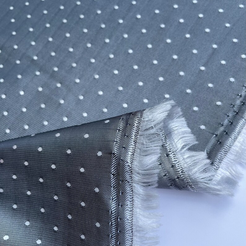 May include: A close-up shot of grey fabric featuring white polka dots. The fabric has a textured surface and a fringed edge. The fabric is folded, showing the underside with a striped pattern.