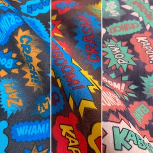 May include: A colourful fabric with a comic book theme. The fabric features various words in a bold, graphic font, including "CRASH!" and "WHAM!" The fabric is a mix of blue, red, yellow, green, and black.