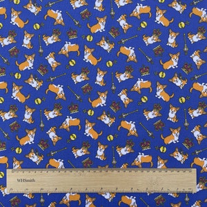 May include: Blue fabric with a repeating pattern of orange and white corgi dogs wearing crowns and holding scepters. The fabric also has a repeating pattern of yellow balls and gold crowns. The fabric is measured with a ruler that says "WHSmith".