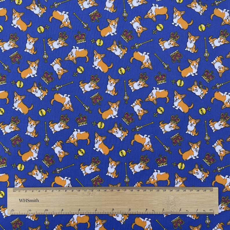 May include: Blue fabric with a repeating pattern of orange and white corgi dogs wearing crowns and holding scepters. The fabric also has a repeating pattern of yellow balls and gold crowns. The fabric is measured with a ruler that says "WHSmith".