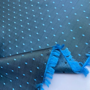 May include: A close-up of a teal fabric with a subtle polka dot pattern in a lighter shade of blue. The fabric has a textured appearance and a fringed edge in a vibrant blue colour. The fabric is folded over.
