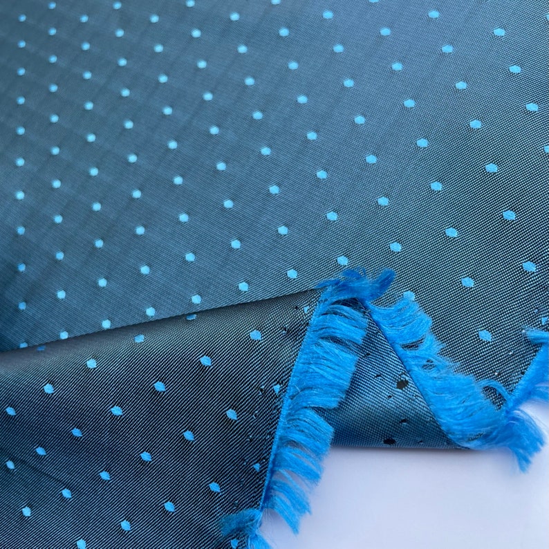 May include: A close-up of a teal fabric with a subtle polka dot pattern in a lighter shade of blue. The fabric has a textured appearance and a fringed edge in a vibrant blue colour. The fabric is folded over.