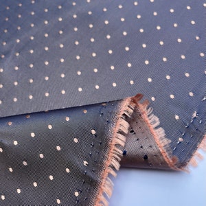May include: A close-up shot of a blue fabric featuring a repeating pattern of small, light-coloured polka dots. The fabric has a subtle sheen and a frayed edge in a contrasting peach colour, showing the layers of the material.