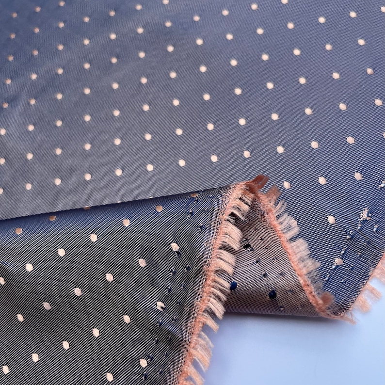 May include: A close-up shot of a blue fabric featuring a repeating pattern of small, light-coloured polka dots. The fabric has a subtle sheen and a frayed edge in a contrasting peach colour, showing the layers of the material.