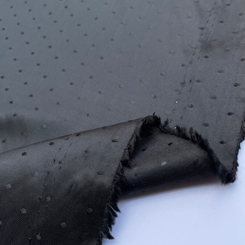 May include: A close-up shot of black fabric with a dotted design. The fabric is folded, showing a raw, frayed edge. The material has a subtle sheen, and the dots are uniformly distributed across the surface.