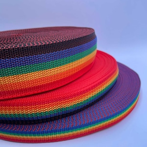 Può includere: Tre rotoli di fettuccia a righe arcobaleno, ognuno con una base di colore diversa: rosso, viola e nero. La fettuccia è tessuta con una trama fine e ha un aspetto brillante e colorato.