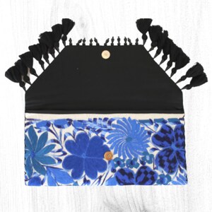 May include: A blue and white floral embroidered clutch with black tassels. The clutch has a black fabric lining and a gold button closure.