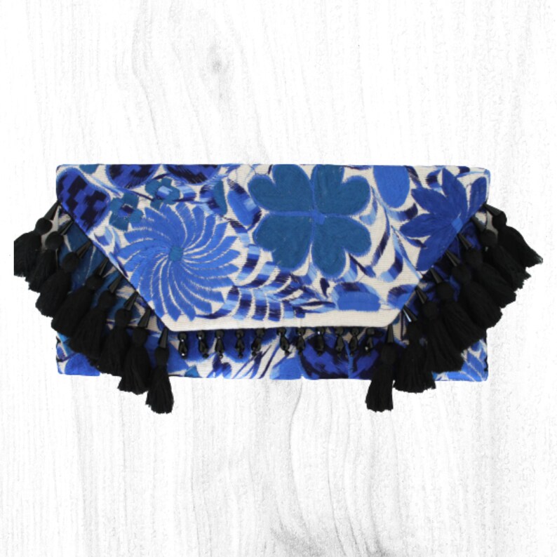 May include: Blue and white embroidered clutch with black tassel trim. The clutch has a beaded detail along the bottom edge.