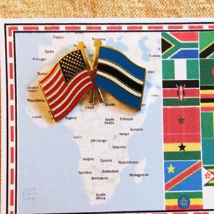 May include: A white card with a red border and a map of Africa on the left side. On the right side is a grid of flags from African countries. Two small pins with the American and Botswana flags are attached to the map.