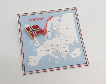 May include: A map of Europe with the country of Norway highlighted in red. A small gold pin with the Norwegian flag is placed on the map.