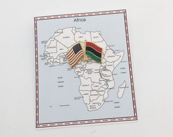 May include: A gold-colored pin with the American and African flags side-by-side, placed on a map of Africa. The map shows the names of countries and bodies of water.