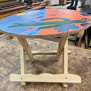 May include: A round wooden folding table with a painted tabletop featuring a desert landscape. The design includes a bright yellow sun, blue mountains, orange ground, and green saguaro cacti with blue flowering succulents on the surface.