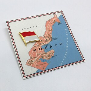 May include: A red and white flag pin with a gold border, placed on a map of Monaco. The map shows the surrounding area of France and the Mediterranean Sea.