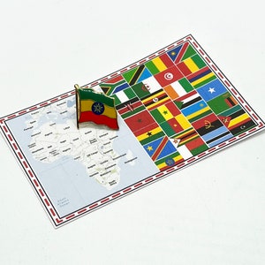 May include: A pin featuring the Ethiopian flag, in red, yellow, and green, is placed on a map of Africa. The pin is next to a card displaying various national flags. The map and flag card are on a white surface.