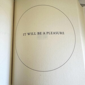 May include: Open book pages with the text "IT WILL BE A PLEASURE" inside a black circle. The text is in a serif font. The pages are off-white.