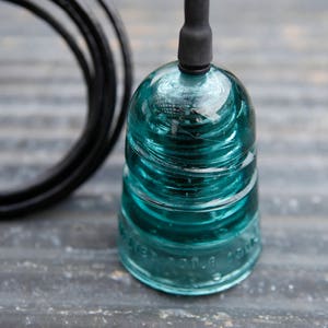 K&ouml;nnte beinhalten: Ein t&uuml;rkisfarbener Glasisolator mit einem daran befestigten schwarzen Draht. Der Isolator hat eine abgerundete Oberseite und eine zylindrische Basis. Der Draht ist um die Basis des Isolators gewickelt.