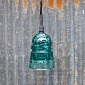 K&ouml;nnte beinhalten: Eine tealblaue Glasisolatoren-Pendelleuchte, die an einem schwarzen Kabel vor einem rostigen Metallhintergrund h&auml;ngt.