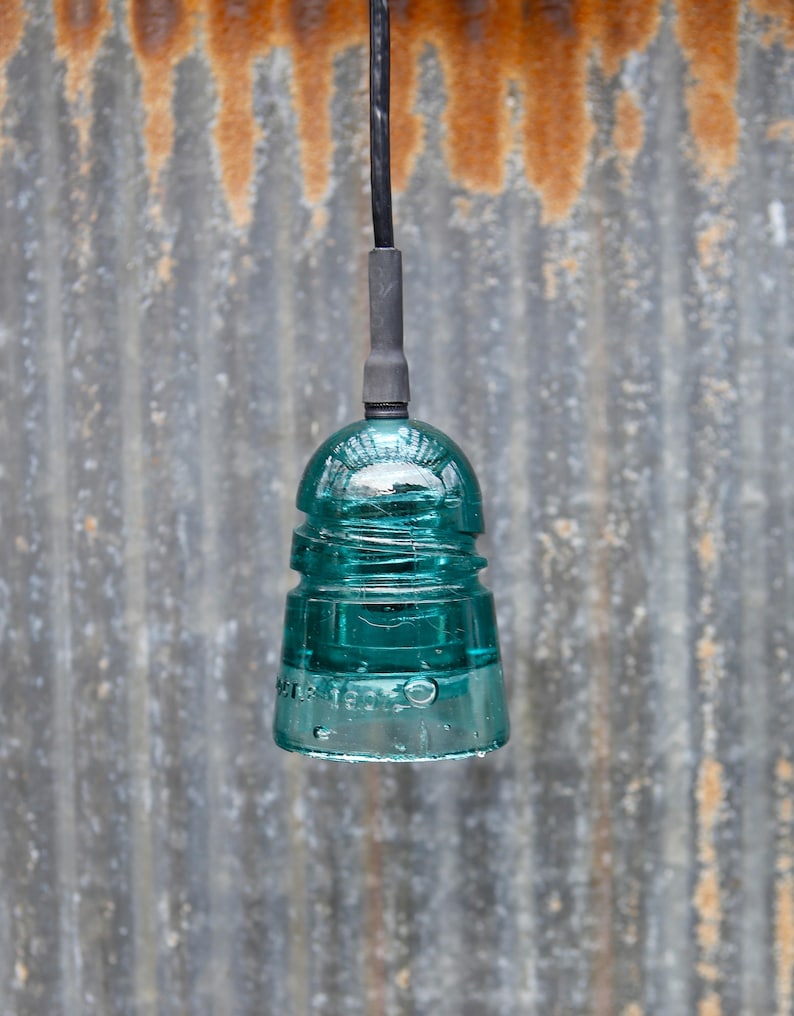 K&ouml;nnte beinhalten: Eine tealblaue Glasisolatoren-Pendelleuchte, die an einem schwarzen Kabel vor einem rostigen Metallhintergrund h&auml;ngt.