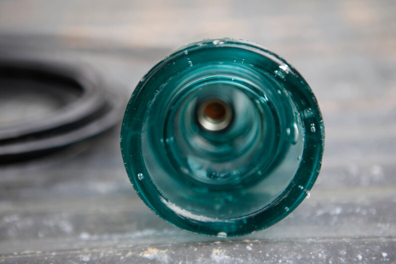 K&ouml;nnte beinhalten: Nahaufnahme eines t&uuml;rkisfarbenen Glasisolatoren mit einem Loch in der Mitte. Der Isolator hat eine glatte, abgerundete Oberfl&auml;che und scheint aus dickem Glas zu sein.