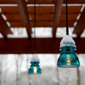 Peut inclure: Trois lampes suspendues en verre bleu avec de la neige sur le dessus, suspendues &agrave; une pergola en bois. Les lumi&egrave;res sont allum&eacute;es et diffusent une lueur chaude.