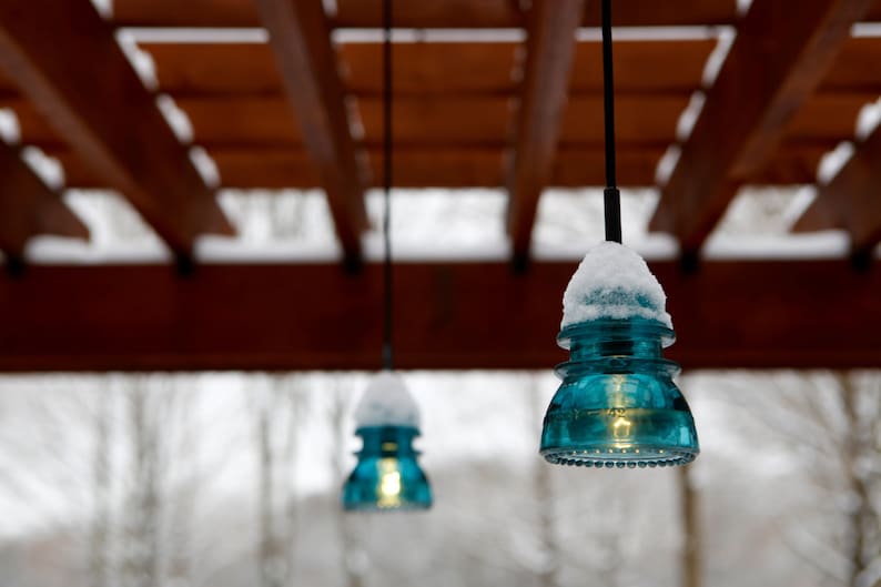 Peut inclure: Trois lampes suspendues en verre bleu avec de la neige sur le dessus, suspendues &agrave; une pergola en bois. Les lumi&egrave;res sont allum&eacute;es et diffusent une lueur chaude.