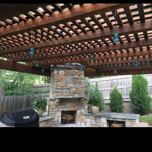 Peut inclure: Une chemin&eacute;e en pierre avec un foyer &agrave; bois et une pergola en bois au-dessus. La pergola a un design en treillis et est d&eacute;cor&eacute;e de lampes suspendues en verre bleu.