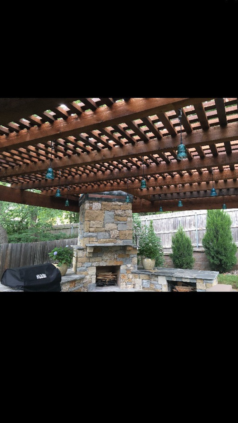 Peut inclure: Une chemin&eacute;e en pierre avec un foyer &agrave; bois et une pergola en bois au-dessus. La pergola a un design en treillis et est d&eacute;cor&eacute;e de lampes suspendues en verre bleu.