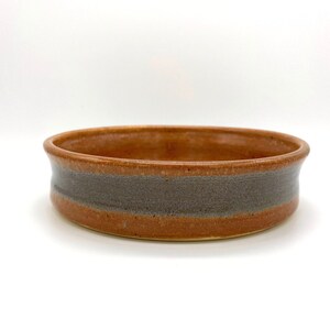 May include: A handmade ceramic bowl with a wide, shallow design. The bowl features a band of gray around the middle, contrasting with the warm brown of the rest of the piece. The bowl is perfect for serving or display.
