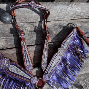 Puede incluir: Un conjunto de brida y peto de cuero marrón para caballos. La brida presenta detalles morados y plateados con joyas decorativas. El peto tiene flecos morados y adornos a juego. El conjunto está diseñado para uso ecuestre.